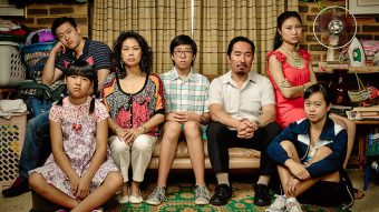 A Chinese family of seven sits on the couch in a suburban home. They are posing as though for a family photo, but are clearly mid-conflict.