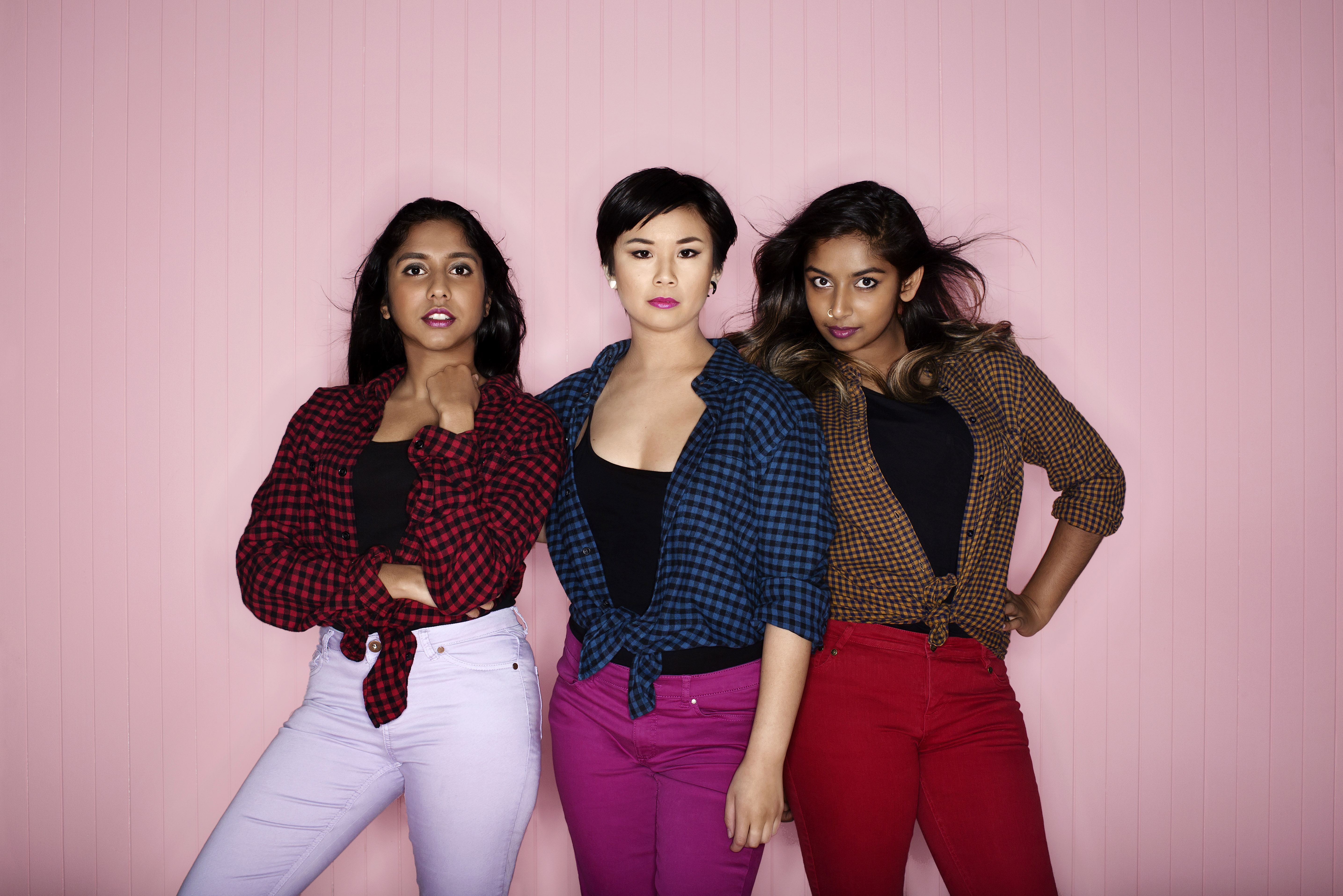 Three young women wearing matching flannel shirts, bright pink lipstick and brightly coloured jeans stand against a pale pink wall. They are posing and staring defiantly at the camera as their hair is blown back by a breeze or fan.