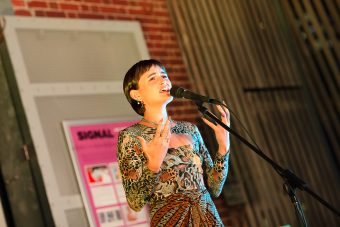 A woman (Komang Rosie Clynes) speaking expressively into a microphone.