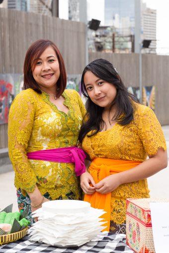 Two women in kebaya standing at a snack table, smiling. 